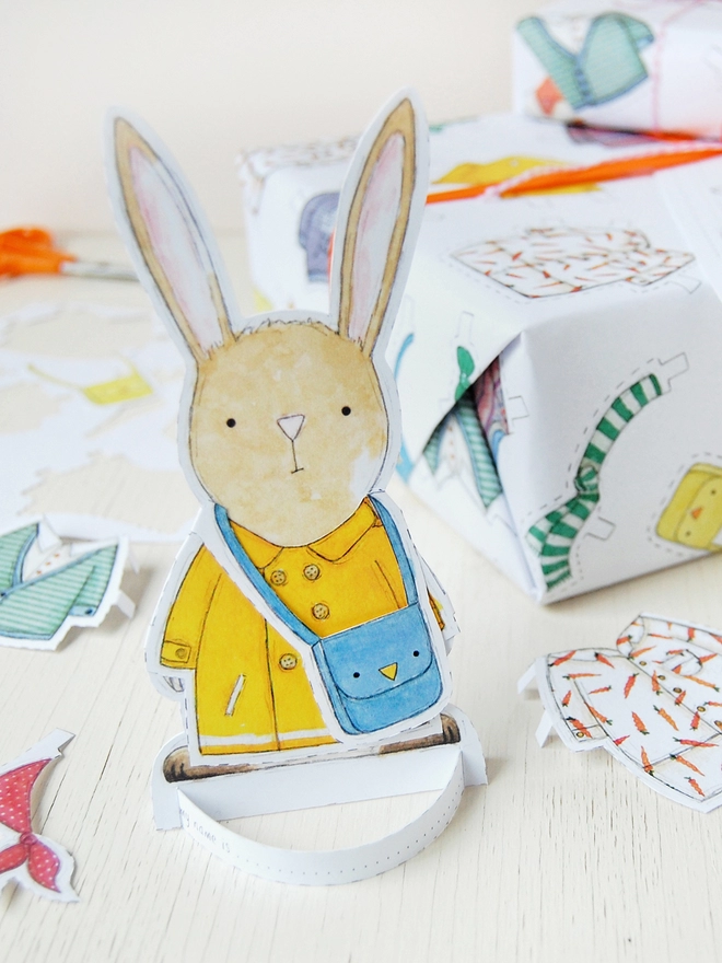 A gift wrapped in white wrapping paper with a hand illustrated outfit design and rabbit paper doll gift tags is beside a cut out rabbit paper doll on a white wooden table.