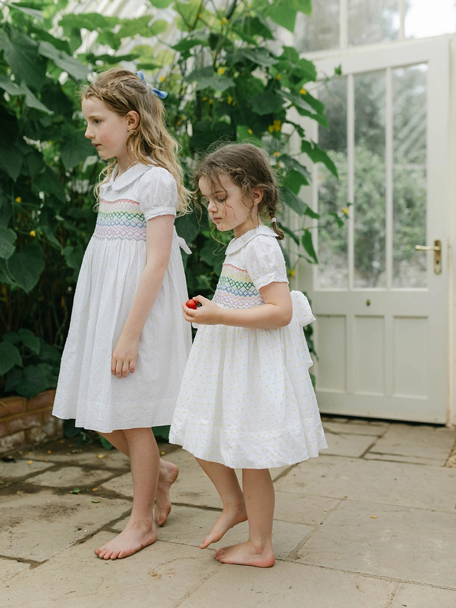 Two girls in a greenhouse both wearing white dresses with smocked rainbow detailing