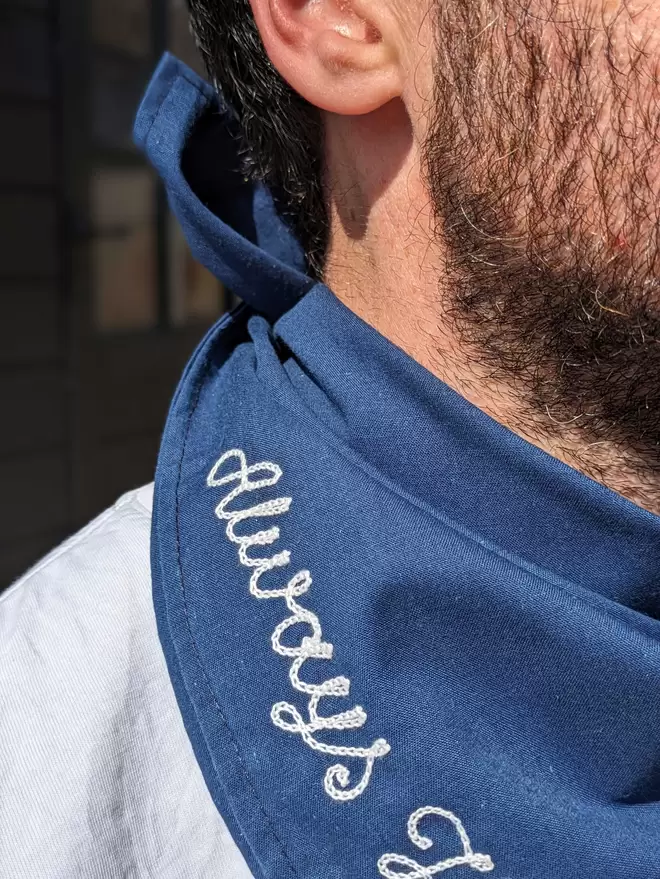 Man wearing a blue Bandana Neckerchief with White Embroidered lettering readiing "Always Hungry, Never Full" close up detail 