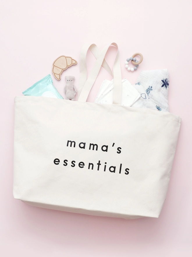 oversized natural canvas tote bag with mama's essentials slogan laying on a pink background with baby essentials spilling out 