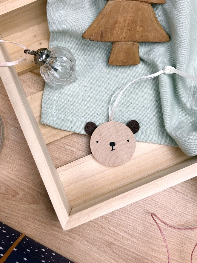 A small wooden and felt bear decoration lays in a wooden crate alongside other Christmas decorations.