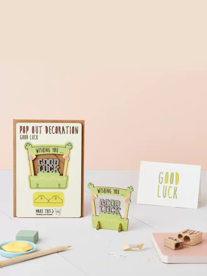 Good luck decoration and good luck card and brown kraft envelope on top of a wooden desk in front of a peachy pink coloured background