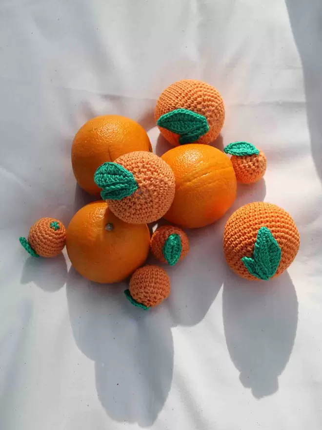 Three crochet oranges and three regular oranges arranged on a soft white background.