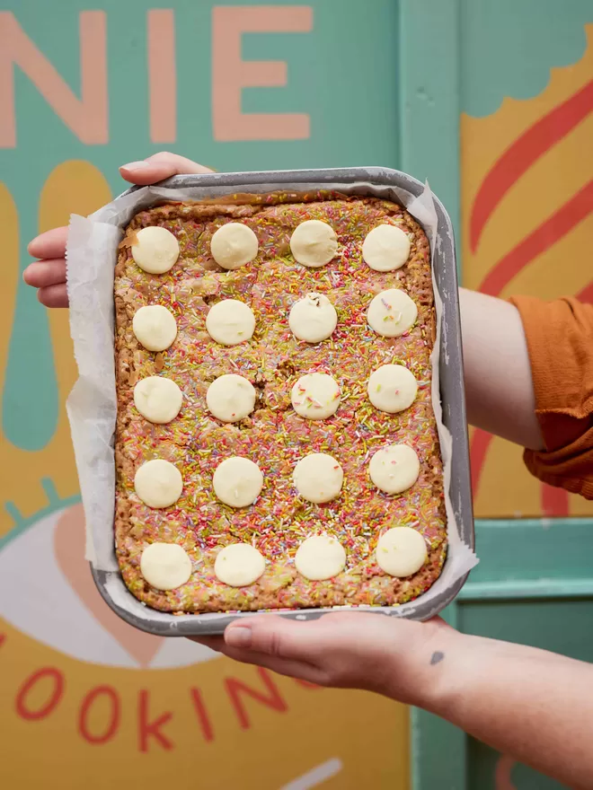 Freshly baked Funfetti blondies with sprinkles and white chocolate buttons being held in a baking tray against a colourful background