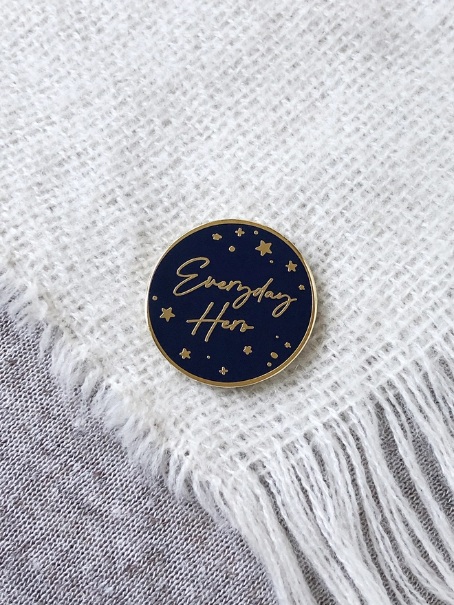 A navy blue and gold enamel pin badge with a gold star design and the words "Everyday Hero" is pinned to a white scarf.
