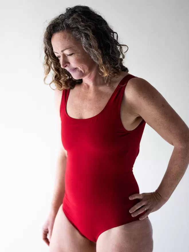 Woman with brown curly hair in studio looking downwards with hand on hip wearing Davy J Sustainable Waterwear red classic crossback swimsuit
