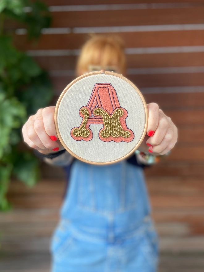 An embroidered letter A in peach with gold beads in a hoop 
