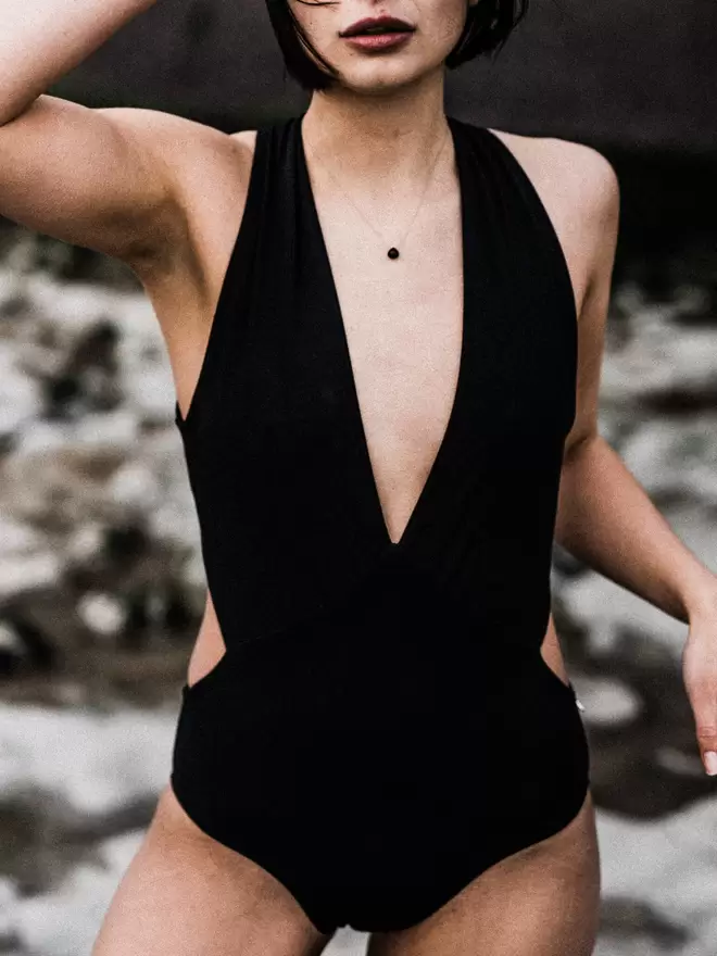 Woman stood on beach frowning with one hand on head, wearing black Davy J Sustainable Waterwear cutout swimsuit with plunging neckline