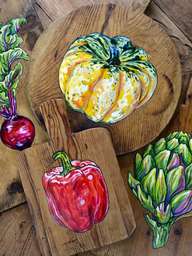 A collection of wooden chopping boards with different handpainted designs including a squash, an artichoke, a beetroot and a red pepper