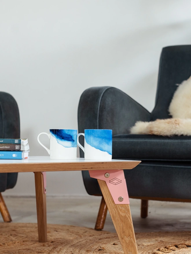 side view of a stylish and minimalist low coffee table with white Fenix top, pink coloured steel brackets and solid oak legs on a jute rug surrounded by blue chairs, with blue and white tea cups on it
