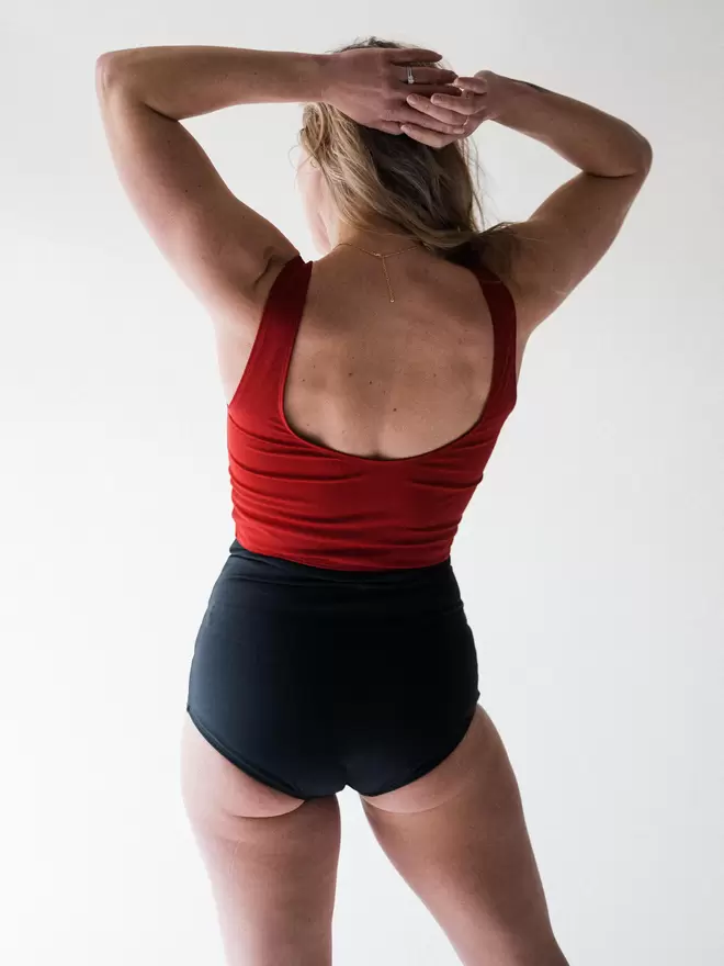 Woman in studio facing away with hands in her blonde hair wearing Davy J Sustainable Waterwear red crop swim top with scoop back overlapping black high waist bikini briefs