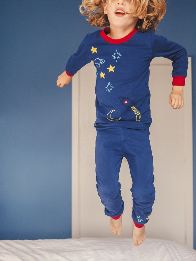 A boy in navy and red pjs with a space scene jumps on a bed