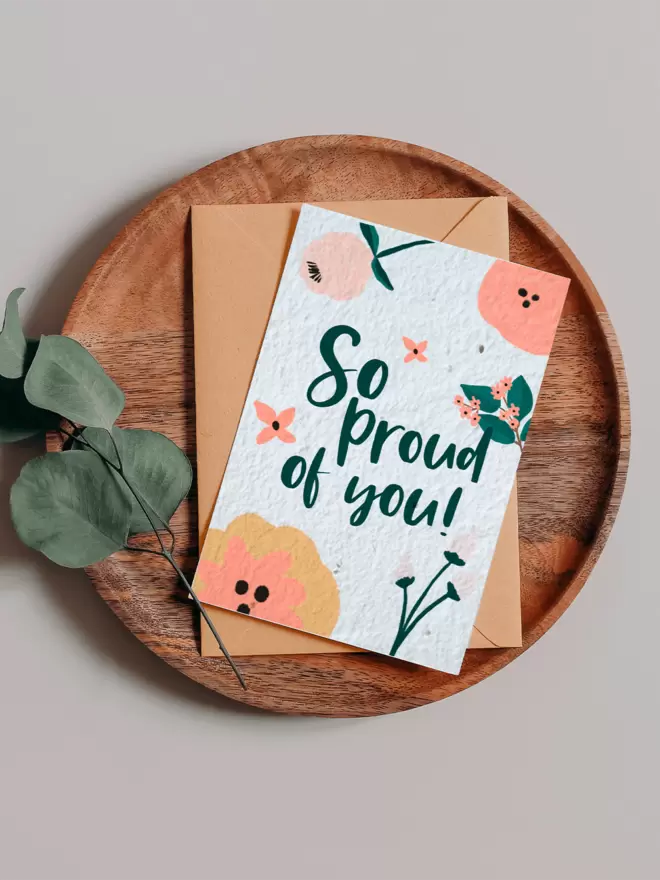 Plantable Card with ‘So Proud of you’ with floral illustrations on a wooden tray next to a Eucalyptus branch