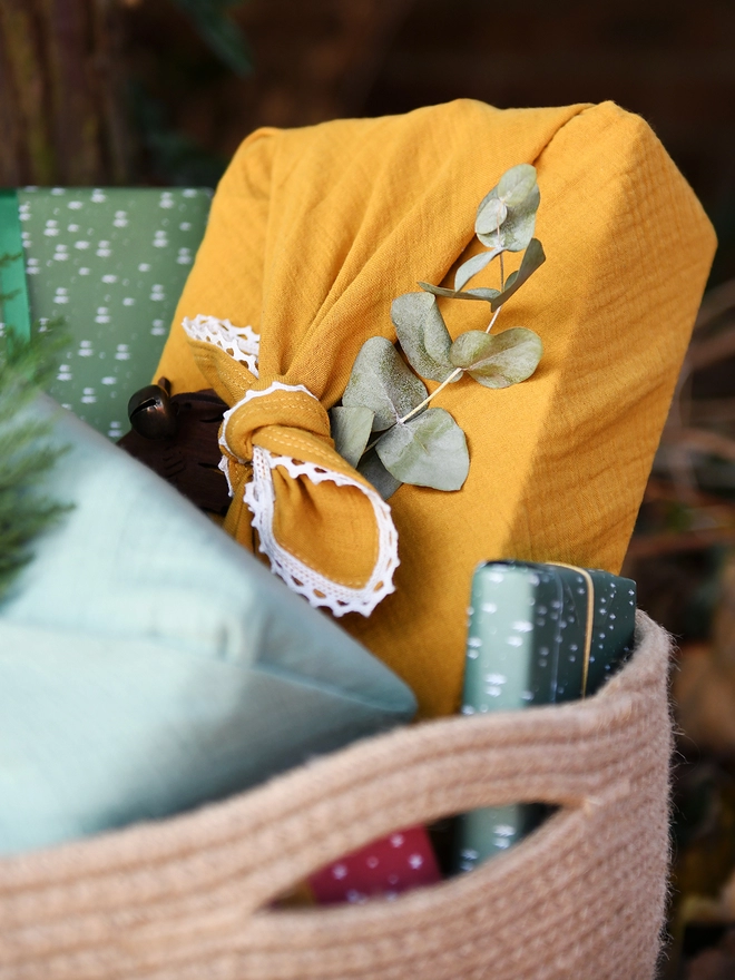 A gift wrapped in mustard yellow cotton fabric wrap with an ivory lace trim is tucked in a woven basket alongside several other wrapped gifts.