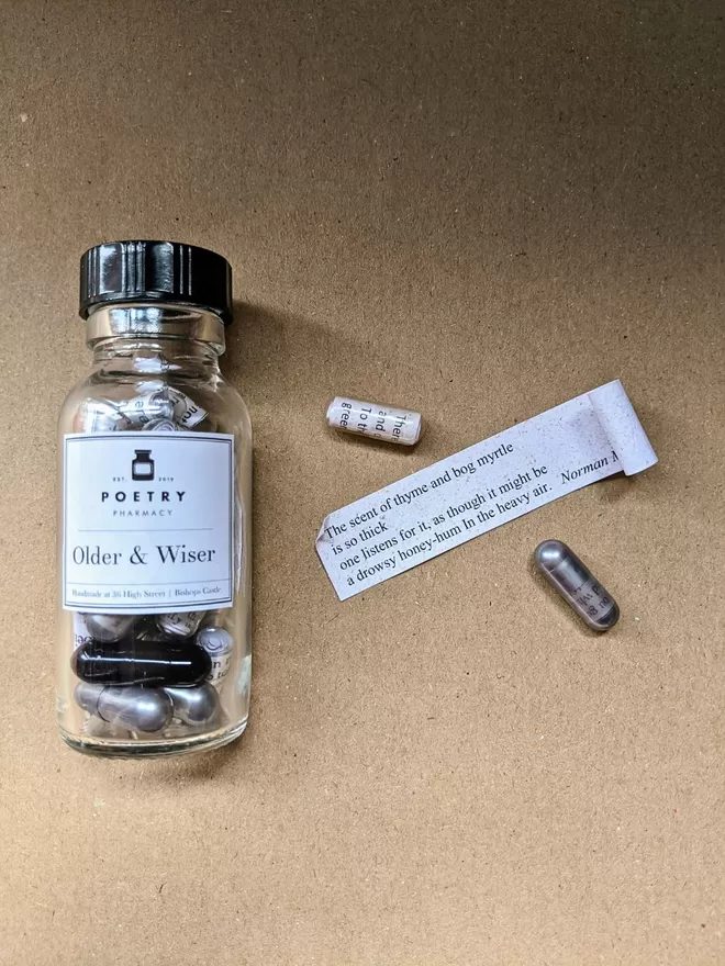 Glass pill bottle containing silver, black and clear Older & Wiser poetry pills printed on banana paper