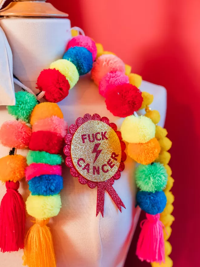 A rosette with a gold disc in the centre featuring the words ‘FUCK CANCER’ and a lightening bolt in red. The scalloped edging and tails are red.