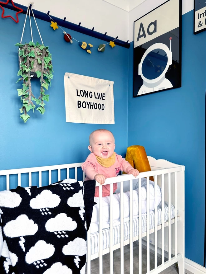 A view of a modern space themed nursery painted a vibrant blue. A baby stands in his cot, holding onto the rail laughing. A black and white monochrome storm cloud blanket is draped over the side. A monochrome print of  ‘A is for Astronaut’ hangs on the wall.