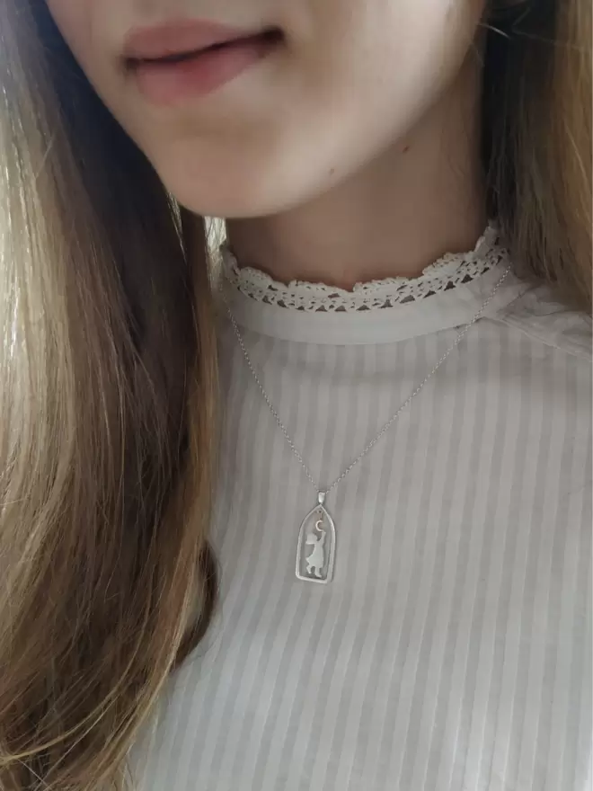 Close up shot of young girl in white high neck shirt and hair flowing down over shoulders, she smiles lightly and suspended on chain around neck is a sterling silver necklace, pendant shows arch shaped frame with silver sillouhete of child reaching up towards a hanging crescent moon that is gold.
