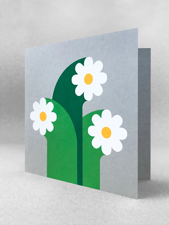 Three white flowers with oversized stems screenprinted on grey square card. Stood in a light grey studio set.