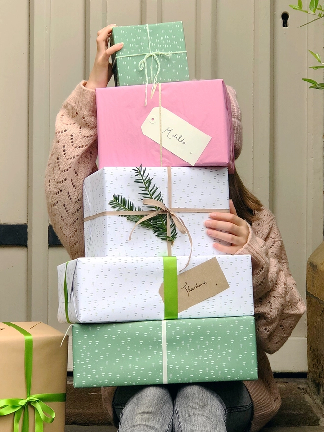 A stack of gifts wrapped in green and white paper with a tiny tree design are being held in front of a door.