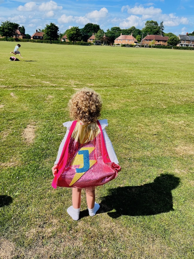 A young girl wearing a hero cape. Her cape has pink sequins, a blue letter 'J' and a gold lightening bolt.