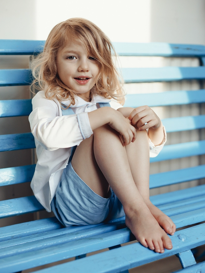 A boy in a white shirt and blue shorts sits on a blue bench
