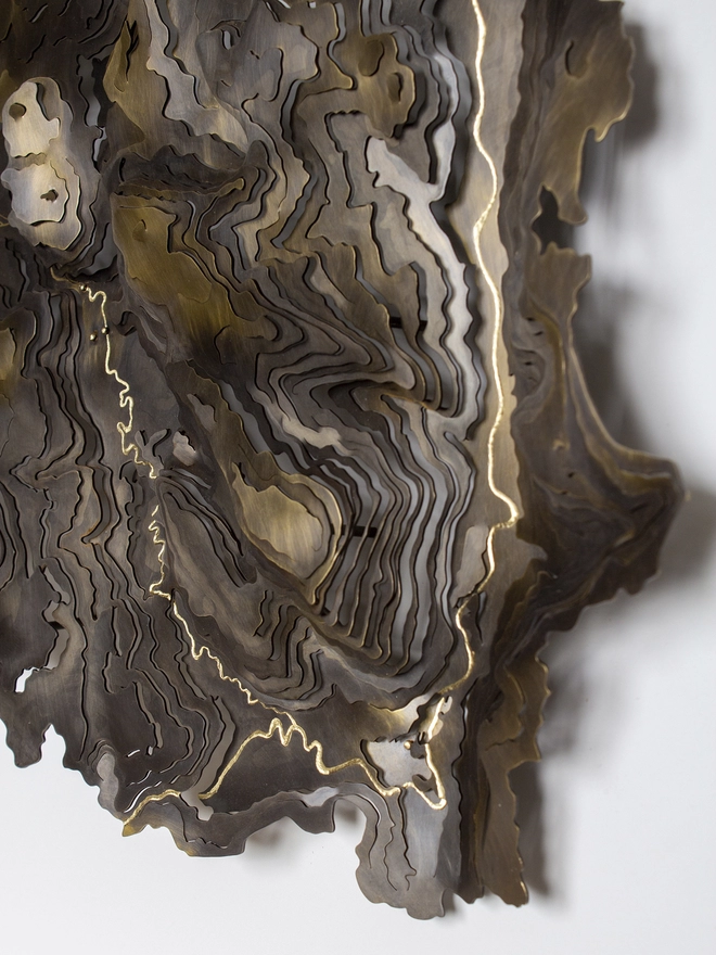 a close up side on shot showing the detail of a metal topographical map, the metal has been blackened and the river engraved showing the gold of the brass.