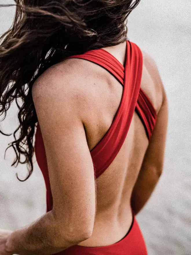 Close up of back of woman with long wavy hair blowing in the wind wearing red Davy J Sustainable Waterwear cutout swimsuit with wide cross back straps