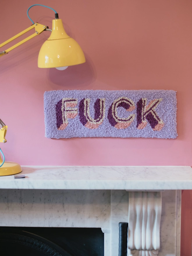 'FUCK' Handmade Tufted Rug/Wall Hanging in purple seen on a pink wall with a yellow lamp.