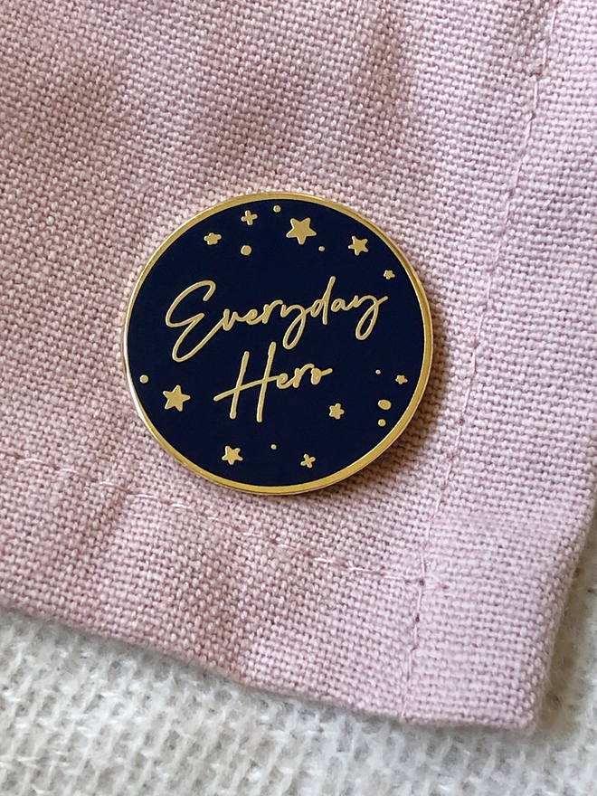 A navy blue and gold enamel pin badge with a gold star design and the words "Everyday Hero" is pinned to a pink blanket.