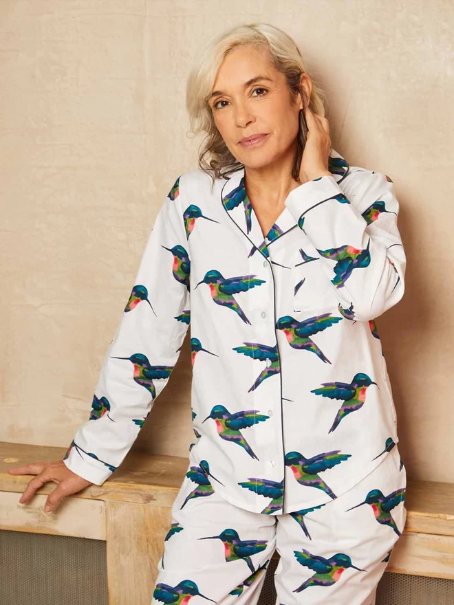 Model has her hand behind her ear looking at the camera while wearing a traditional shirt style pyjama with navy piping on a fresh white based print covered in watercolour hummingbirds