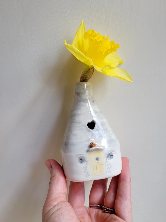 Bud vase house held between fingers and thumb of a hand the house vase has windows and a yellow door with a modelled robin above it and a heart cut out in the grey roof there is a bright yellow daffodil in the flower vase  