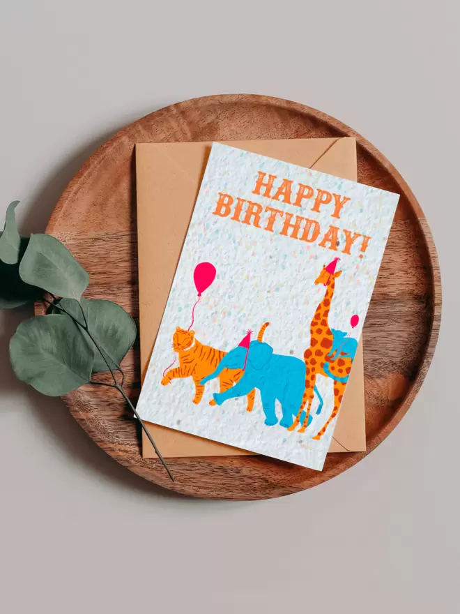 Plantable Card with Happy birthday and an illustration of an elephant and giraffe wearing party hats and a tiger and monkey each carrying a balloon on a wooden tray next to a Eucalyptus branch