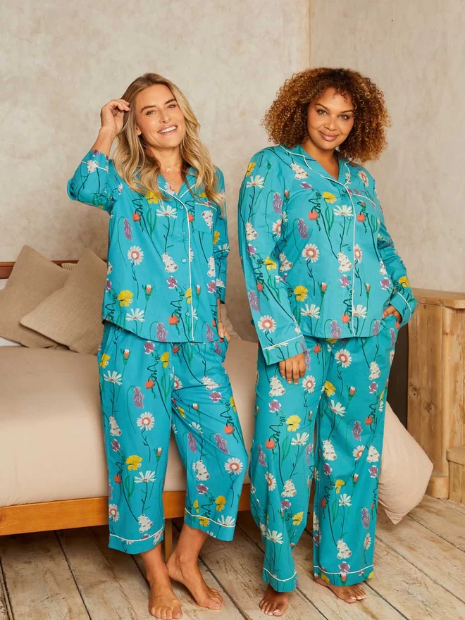 Two models smile out standing in front of a neutral coloured bed, they are wearing traditional style pyjamas with a shirt top and wide legged bottoms in a teal based floral print covered in british wild flowers in a mix of yellows, pinks and purples.