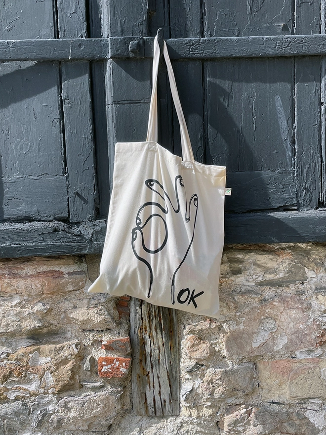  Ok tote bag hanging on old dark blue/grey doors on an old wall. Showing the screenprinted OK design.