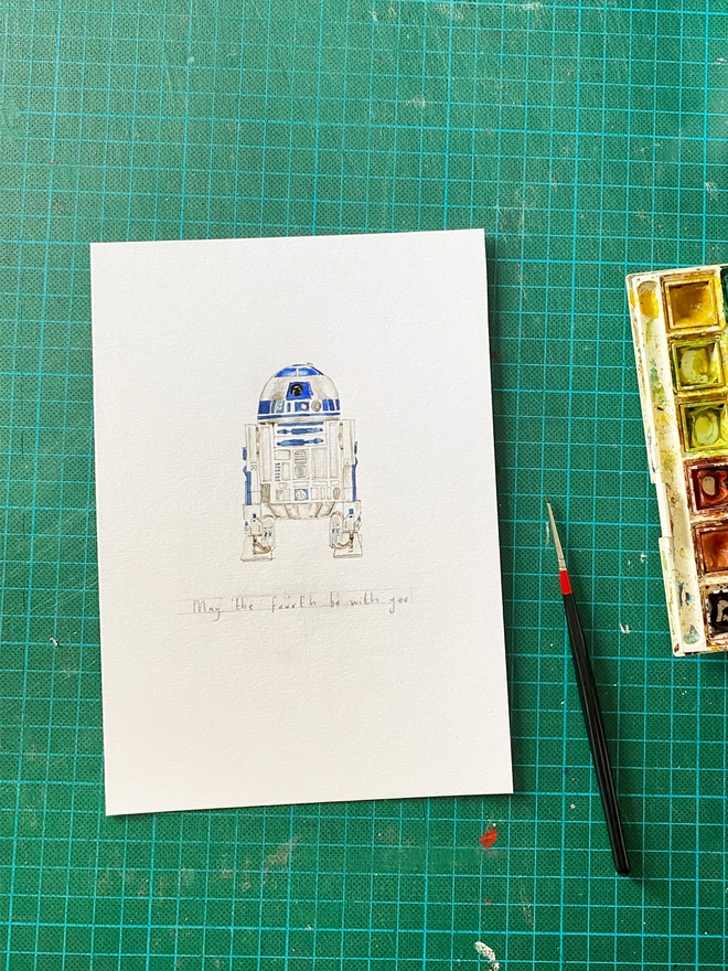 Work in progres photo of a drawn and painted watercolour illustration of R2D2 with a detailed yet organic and slightly loose style. The A5 piece of paper with the watercolour painting on sits on a green cutting mat background with a paint set and brush in view to the right. 