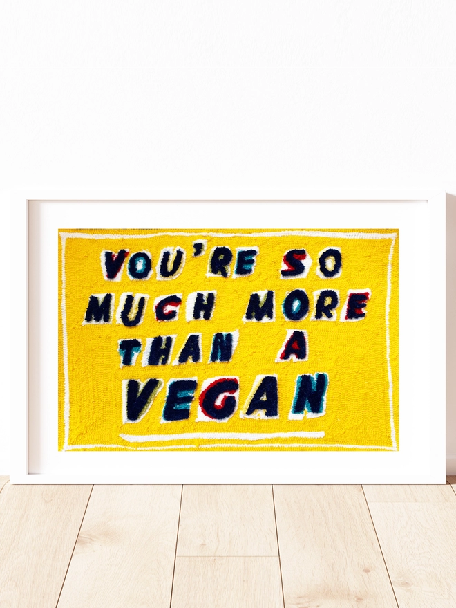 You're so much more than a vegan written in blue tufted lettering with red, blue and yellow tufted shadows on a bright yellow background.  Piece is framed in a white box frame and sat on a wooden floor