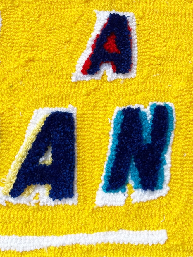 close up of letters A, A, N blue tufted wool with red, yellow and blue shadows and white border on a yellow background