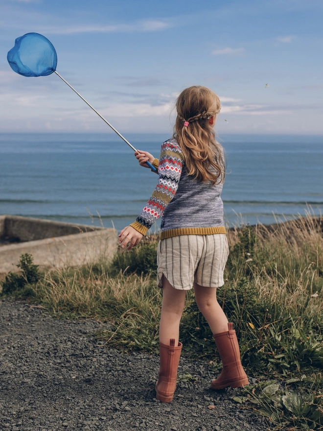 A little girl wearing 'The Daydreamer' Knitted Jumper by The Faraway Gang, holding a blue fishing net near the ocean.