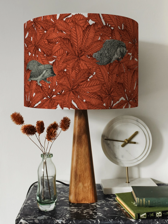 Drum Lampshade featuring hedgehogs in autumnal russet red leaves on a wooden base on a shelf with books and ornaments