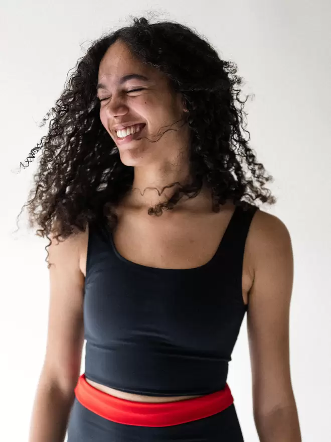 A lady with black curly hair smiling, wearing a Davy J Sustainable Waterwear black cropped swim top with shoulder straps and black high waist briefs with waist rolled down and showing red lining