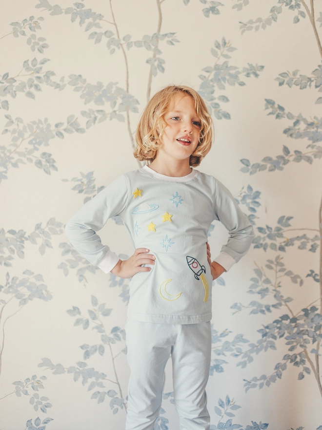 A boy in pale blue grey pyjamas with an embroidered space scene stands with his hands on his hips