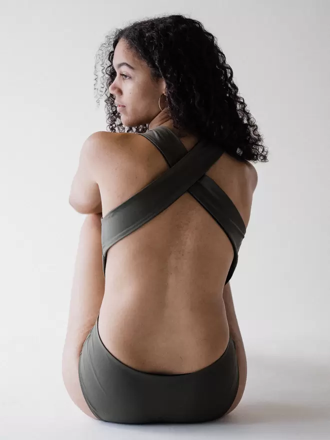  Back view of woman with long curly brown hair sat on the floor in studio wearing an olive green Davy J Sustainable Waterwear cutout swimsuit with wide cross back straps