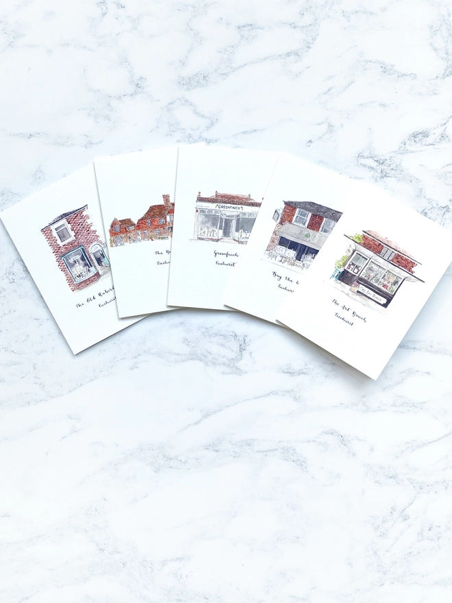 Watercolour illustrated Ticehurst shop front cards on A6 white cards. Featuring five beautiful independent small businesses in Ticehurst, East Sussex - The Bell, Buy the Weigh and The Art Bunch, Greenfinch and The Old Haberdashery. The cards are fanned out on a pale white marble background. 