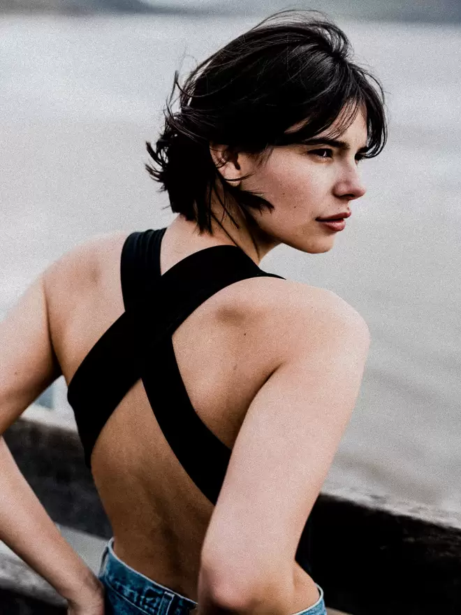 Woman with brunette short hair facing away to the sea, wearing jeans over her black Davy J Sustainable Waterwear cutout swimsuit with wide cross back straps