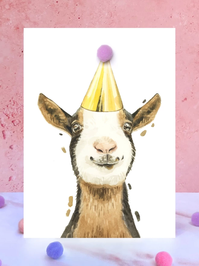 A greeting card featuring a hand painted design of a goat, stood upright on a marble surface surrounded by pompoms. 