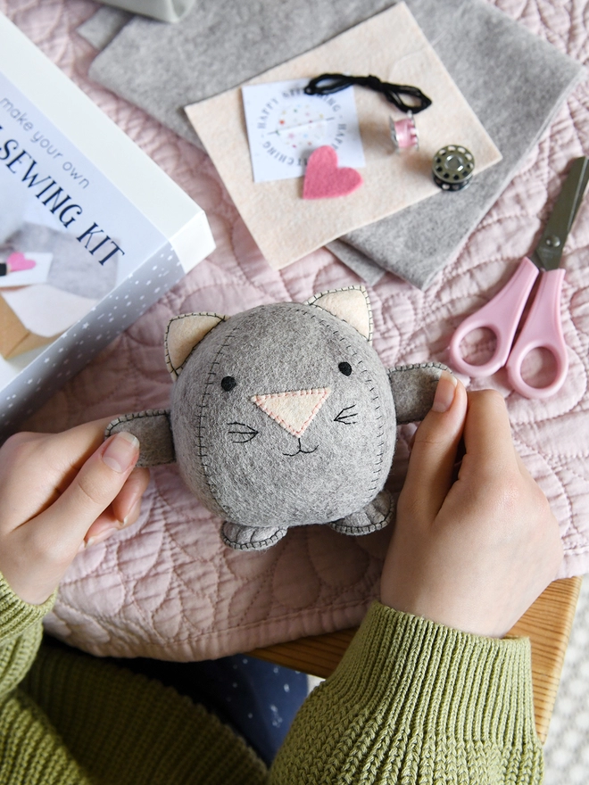 A handmade felt kitten toy is being held in two hands above a desk covered in craft kit components.