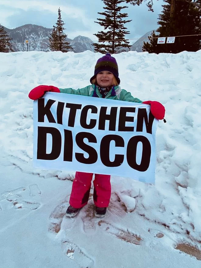 London Drying Kitchen Disco screen printed in black text on white tea towel held by little girl in the snow