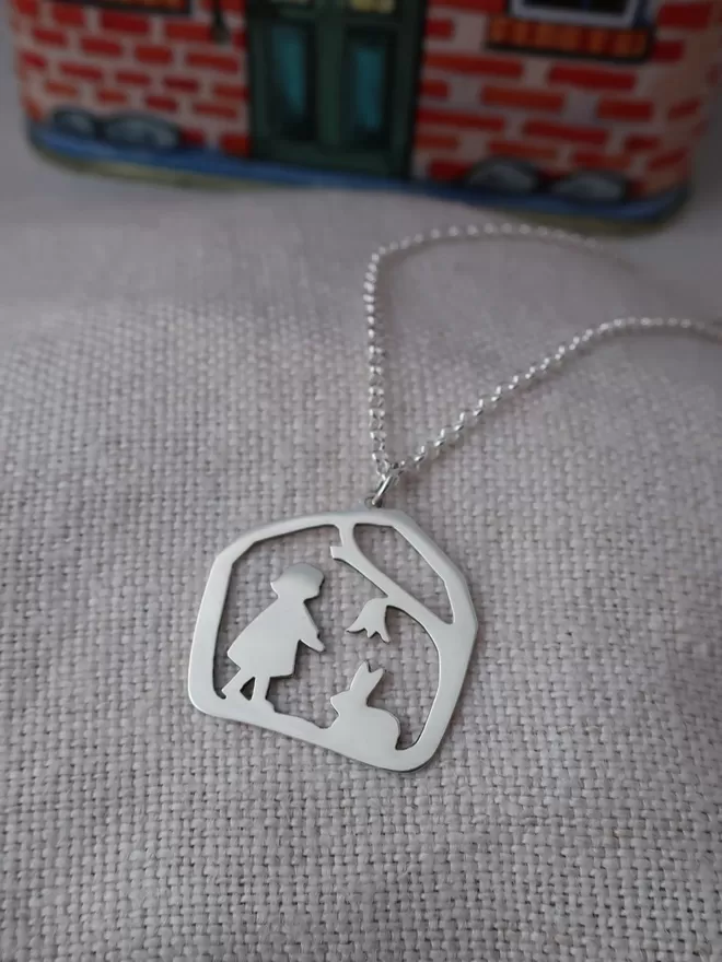 Silver pendant sits on linen, chain leads off into background, pendant shows sillouhette of child reaching towards rabbit that sits at her feet, both stand underneath a tree branch with flower, sterling silver.