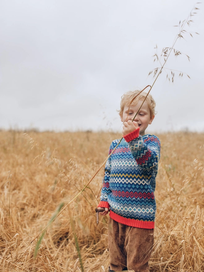 A little boy standing in a field holding some corn wearing 'The Daydreamer' Knitted Jumper from The Faraway Gang.